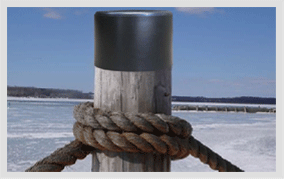 A black heat-shrink cap recovered over the sawn top of a timber dock pile, a mooring rope coiled around the pile below it, with a frozen harbour behind.